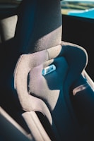 A detailed view of a car seat with textured fabric and a shiny logo in the center. The seat appears ergonomic and is structured for comfort with contours and padding. It is partially illuminated by natural light, suggesting it is in a vehicle exposed to sunlight.
