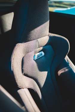A detailed view of a car seat with textured fabric and a shiny logo in the center. The seat appears ergonomic and is structured for comfort with contours and padding. It is partially illuminated by natural light, suggesting it is in a vehicle exposed to sunlight.