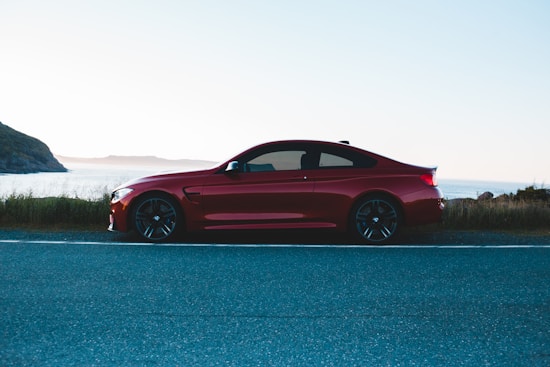 A sleek red sports car is parked on the side of a coastal road. The background features a large body of water, likely an ocean or sea, with hills or cliffs on the left side. The environment seems calm with a clear sky and minimal vegetation along the road.