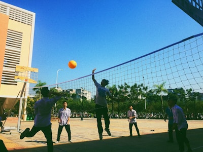 Several people are playing volleyball in an outdoor area. The sky is clear and blue, and there's a net set up between two poles. One player is jumping to hit an orange volleyball, while others are positioned around the net. There is a modern building to the side and some trees in the background.