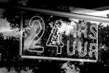 A neon sign displays the text '24 HRS 7 UUR' in bold letters. The background features reflections of trees, likely captured through a reflective surface such as glass.