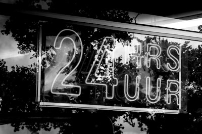 A neon sign displays the text '24 HRS 7 UUR' in bold letters. The background features reflections of trees, likely captured through a reflective surface such as glass.