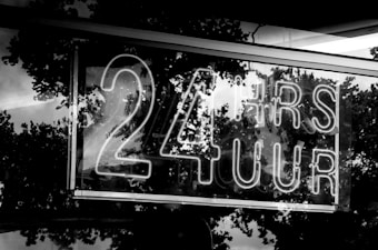A neon sign displays the text '24 HRS 7 UUR' in bold letters. The background features reflections of trees, likely captured through a reflective surface such as glass.