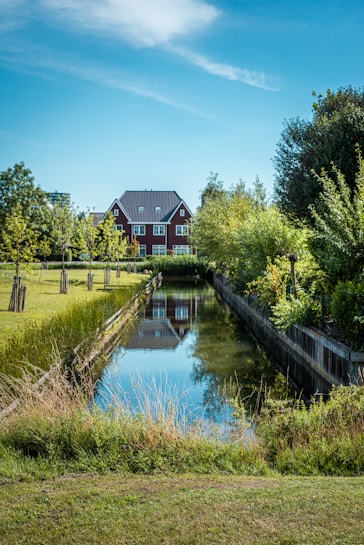 A peaceful, serene landscape with a clear, calm canal reflecting the blue sky and surrounding greenery. The backdrop features a charming red brick house with a dark roof, nestled among lush trees. The grass along the canal is neatly trimmed, contributing to a tidy and picturesque setting.
