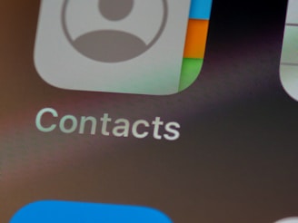 Close-up view of a smartphone screen displaying the Contacts application icon. The icon features a stylized silhouette of a person alongside multicolored gradients. Below the icon, the word 'Contacts' is clearly visible against a gray background.
