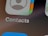 Close-up view of a smartphone screen displaying the Contacts application icon. The icon features a stylized silhouette of a person alongside multicolored gradients. Below the icon, the word 'Contacts' is clearly visible against a gray background.