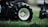 Close-up of a large black tire with a white rim resting on lush green grass. The tire treads are visible, emphasizing its texture and ruggedness. The background is blurred, focusing the attention on the tire and grass.