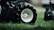 Close-up of a large black tire with a white rim resting on lush green grass. The tire treads are visible, emphasizing its texture and ruggedness. The background is blurred, focusing the attention on the tire and grass.