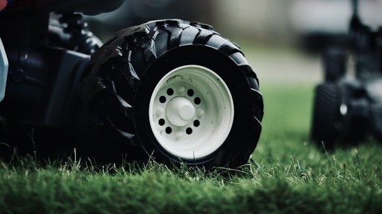 Close-up of a large black tire with a white rim resting on lush green grass. The tire treads are visible, emphasizing its texture and ruggedness. The background is blurred, focusing the attention on the tire and grass.
