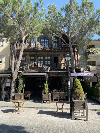 A rustic restaurant with a wooden exterior is located amid tall, leafy trees. The building includes a stone facade with small balconies on the upper floor, and there are outdoor tables and chairs arranged in front. The scene is bathed in sunlight, giving it a warm and inviting atmosphere.
