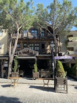 A rustic restaurant with a wooden exterior is located amid tall, leafy trees. The building includes a stone facade with small balconies on the upper floor, and there are outdoor tables and chairs arranged in front. The scene is bathed in sunlight, giving it a warm and inviting atmosphere.