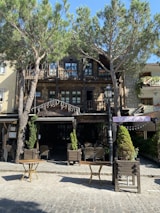 A rustic restaurant with a wooden exterior is located amid tall, leafy trees. The building includes a stone facade with small balconies on the upper floor, and there are outdoor tables and chairs arranged in front. The scene is bathed in sunlight, giving it a warm and inviting atmosphere.