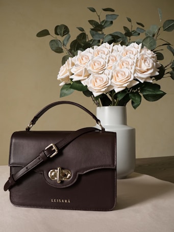 A brown leather handbag with a silver buckle rests on a table. Behind it, a white vase holds a bouquet of cream-colored roses with green foliage. The background is a muted beige color, complementing the elegant and minimalistic setup.