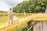 A person wearing safety gear, including a helmet and uniform, is standing on a bright yellow metal walkway. The walkway is elevated with railings and is situated in a lush green area with dense greenery in the background. Overhead street lamps lining the path are visible.
