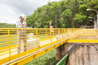 A person wearing safety gear, including a helmet and uniform, is standing on a bright yellow metal walkway. The walkway is elevated with railings and is situated in a lush green area with dense greenery in the background. Overhead street lamps lining the path are visible.