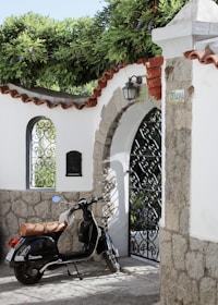 A black scooter is parked in front of a charming stone and whitewashed wall with an ornate metal gate. Above the gate is lush greenery with purple flowers draping over the edge. The scene is shaded by a hanging lantern, and there's a metal mailbox affixed to the wall. The wall features red-tiled accents and a street sign reading 'Giovanna'.