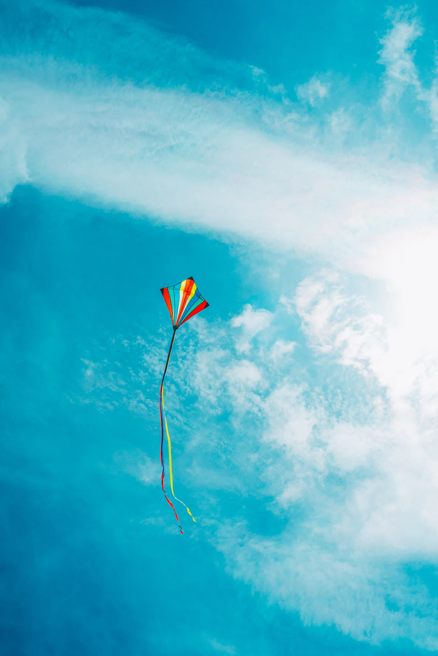 A small rainbow kite with a long tail high in a cloudy blue sky