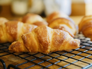Several golden-brown croissants are placed on a black cooling rack. The croissants appear freshly baked, with a flaky and shiny texture. The background is slightly blurred, focusing attention on the pastries.
