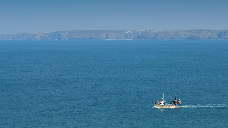 A single small fishing boat crossing open blue water far below a distant line of coastal cliffs