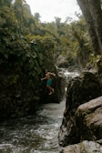 A person is mid-air in a leap over a narrow stream with lush green vegetation and rocky formations surrounding the area. The water appears to be flowing through a small gorge under a canopy of dense trees. The scene reflects a sense of adventure and exploration.
