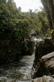 A person is mid-air in a leap over a narrow stream with lush green vegetation and rocky formations surrounding the area. The water appears to be flowing through a small gorge under a canopy of dense trees. The scene reflects a sense of adventure and exploration.