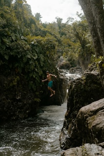 A person is mid-air in a leap over a narrow stream with lush green vegetation and rocky formations surrounding the area. The water appears to be flowing through a small gorge under a canopy of dense trees. The scene reflects a sense of adventure and exploration.