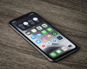 A smartphone with a black frame lies on a wooden surface, displaying various app icons on its screen such as clocks, settings, photos, and social media applications. The home screen has a dark-themed background, and the time shows 2:14.