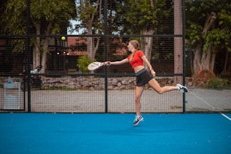 A person in athletic wear is playing a racquet sport on a bright blue court. The player is mid-motion, preparing to hit a ball with the racquet. In the background, there is a wire fence, trees, and some urban structures.