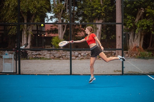 A person in athletic wear is playing a racquet sport on a bright blue court. The player is mid-motion, preparing to hit a ball with the racquet. In the background, there is a wire fence, trees, and some urban structures.