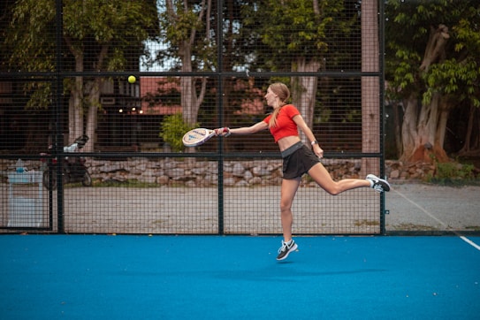 A person in athletic wear is playing a racquet sport on a bright blue court. The player is mid-motion, preparing to hit a ball with the racquet. In the background, there is a wire fence, trees, and some urban structures.