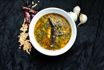 A bowl of soup featuring a dark brown chili pepper and leafy greens is placed on a black surface. Surrounding the bowl are scattered dried split peas, dried red chilies, and whole garlic bulbs.
