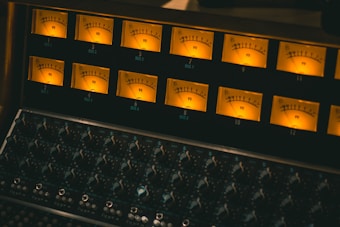 A close-up view of a vintage audio mixing console with multiple illuminated VU meters and numerous dials and knobs beneath them. The warm glow of the meters creates a retro and technical atmosphere.