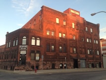 A large red-brick building with multiple stories is depicted, featuring numerous rectangular and arched windows. The setting sun casts a warm glow on the facade, accentuating its architectural details. A sign on the building advertises personal fitness training. The street in front of the building is mostly empty, with a fire hydrant and streetlight visible.