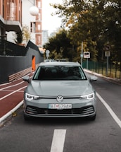 A silver Volkswagen car is parked on a city street, lined with trees and modern buildings. The foreground shows a clear view of the vehicle from the front, with distinct license plates. The street features a bike lane and various traffic signs, and the background includes greenery and urban structures.
