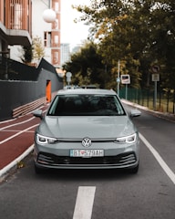 A silver Volkswagen car is parked on a city street, lined with trees and modern buildings. The foreground shows a clear view of the vehicle from the front, with distinct license plates. The street features a bike lane and various traffic signs, and the background includes greenery and urban structures.
