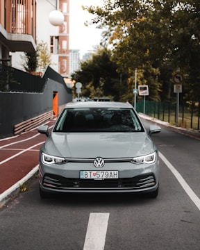 A silver Volkswagen car is parked on a city street, lined with trees and modern buildings. The foreground shows a clear view of the vehicle from the front, with distinct license plates. The street features a bike lane and various traffic signs, and the background includes greenery and urban structures.