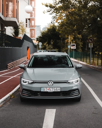 A silver Volkswagen car is parked on a city street, lined with trees and modern buildings. The foreground shows a clear view of the vehicle from the front, with distinct license plates. The street features a bike lane and various traffic signs, and the background includes greenery and urban structures.