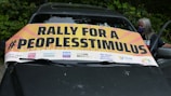 A banner with bold text reads 'Rally for a #PeoplesStimulus' positioned on the windshield of a car. Logos for various advocacy groups and initiatives are displayed along the bottom of the banner. A person wearing a mask stands beside the car against a backdrop of lush green foliage.