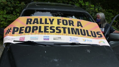 A banner with bold text reads 'Rally for a #PeoplesStimulus' positioned on the windshield of a car. Logos for various advocacy groups and initiatives are displayed along the bottom of the banner. A person wearing a mask stands beside the car against a backdrop of lush green foliage.