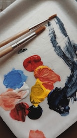 A paint palette holds various dabs of paint in vibrant colors including blue, red, yellow, black, white, and orange. Two paintbrushes with wooden handles and metal ferrules rest on the palette, with some paint smeared alongside them.