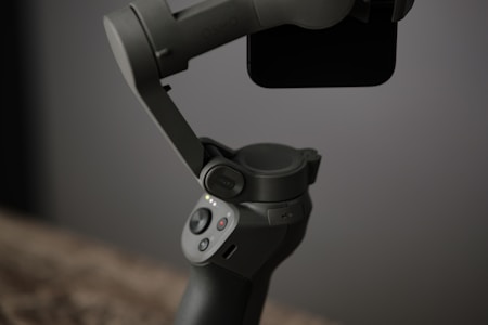 A handheld gimbal stabilizer designed for smartphones is prominently featured. The device has a sleek, matte finish with a small control panel featuring buttons and indicator lights. It is partially holding a smartphone in place.