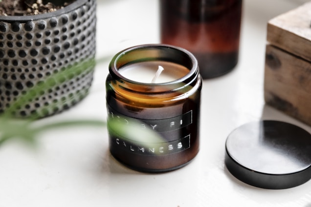 A small amber glass candle jar with a white wick is placed on a white surface. Nearby, there's a black lid resting on the surface, and a textured plant pot can be seen in the background. A wooden box is partially visible on the right side.