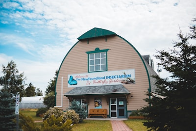 A large wooden building with a curved roof and teal trim. The building features a prominent sign reading 'The Newfoundland Insectarium & Butterfly Garden.' The entrance is flanked by small bushes and benches. The surrounding area is landscaped with trees and shrubs, beneath a partially cloudy sky.