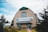 A large wooden building with a curved roof and teal trim. The building features a prominent sign reading 'The Newfoundland Insectarium & Butterfly Garden.' The entrance is flanked by small bushes and benches. The surrounding area is landscaped with trees and shrubs, beneath a partially cloudy sky.