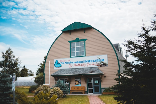 A large wooden building with a curved roof and teal trim. The building features a prominent sign reading 'The Newfoundland Insectarium & Butterfly Garden.' The entrance is flanked by small bushes and benches. The surrounding area is landscaped with trees and shrubs, beneath a partially cloudy sky.
