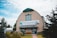 A large wooden building with a curved roof and teal trim. The building features a prominent sign reading 'The Newfoundland Insectarium & Butterfly Garden.' The entrance is flanked by small bushes and benches. The surrounding area is landscaped with trees and shrubs, beneath a partially cloudy sky.
