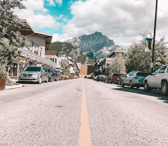 A street view of a small town with a clear road lined with parked cars on both sides. The street is bordered by buildings with various facades, and there are trees and greenery along the sidewalks. In the background, there is a large mountain range under a partly cloudy sky.