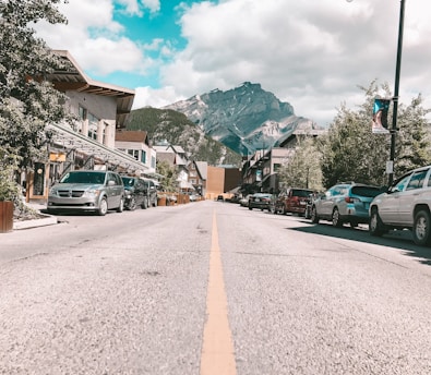 A street view of a small town with a clear road lined with parked cars on both sides. The street is bordered by buildings with various facades, and there are trees and greenery along the sidewalks. In the background, there is a large mountain range under a partly cloudy sky.
