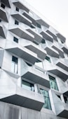 The image features a modern building facade with an intriguing geometric design. The structure is composed of numerous angular, protruding elements that cast subtle shadows. The facade is predominantly gray with glass windows interspersed among the geometric sections, reflecting a bit of light. The architectural style is contemporary and innovative, suggesting a focus on both aesthetics and functionality.