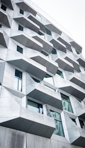 The image features a modern building facade with an intriguing geometric design. The structure is composed of numerous angular, protruding elements that cast subtle shadows. The facade is predominantly gray with glass windows interspersed among the geometric sections, reflecting a bit of light. The architectural style is contemporary and innovative, suggesting a focus on both aesthetics and functionality.